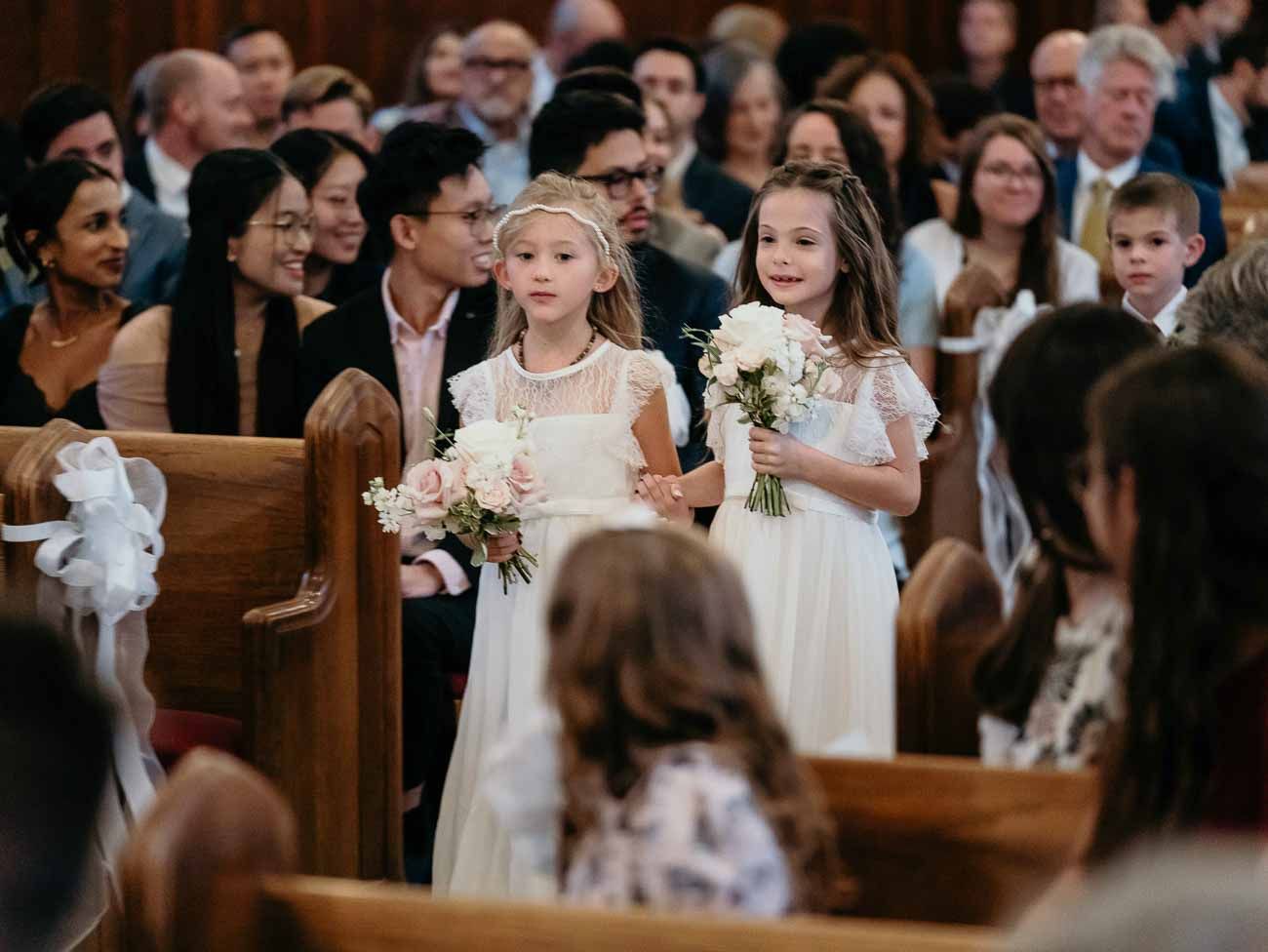 Two flower girls in white dresses walking down wedding aisle.