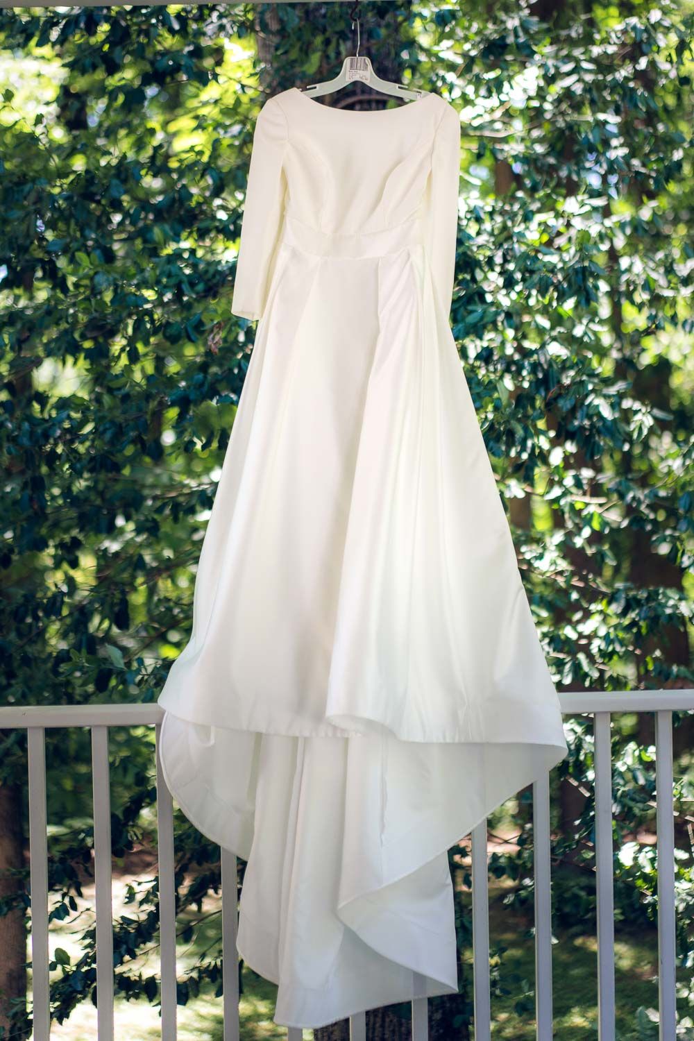 Elegant long-sleeve white wedding gown hanging on balcony railing surrounded by lush green trees.