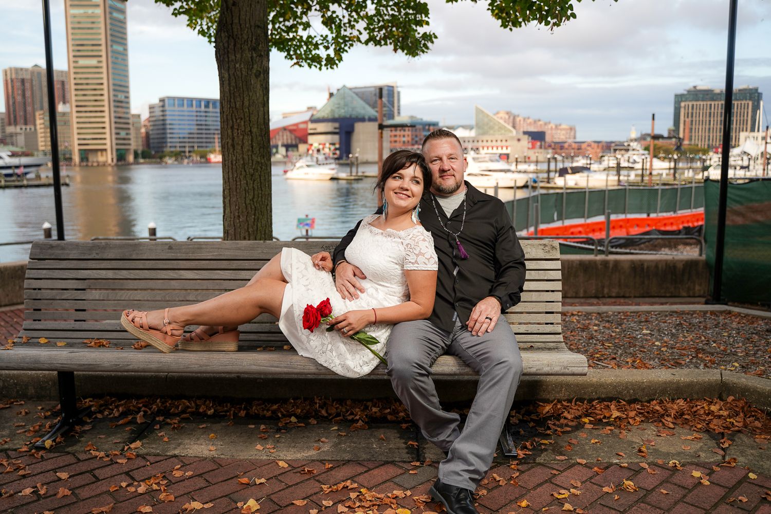 Wedding couple sitting on bench at Baltimore Inner Harbor, bride in lace dress with red rose.