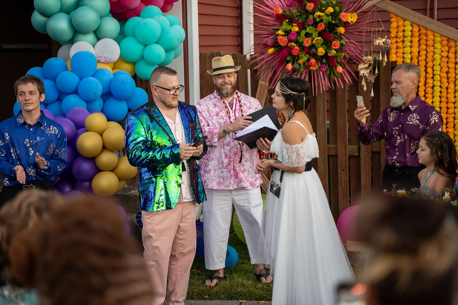 Wedding party stands at colorful ceremony altar with rainbow balloons as officiant reads from book.