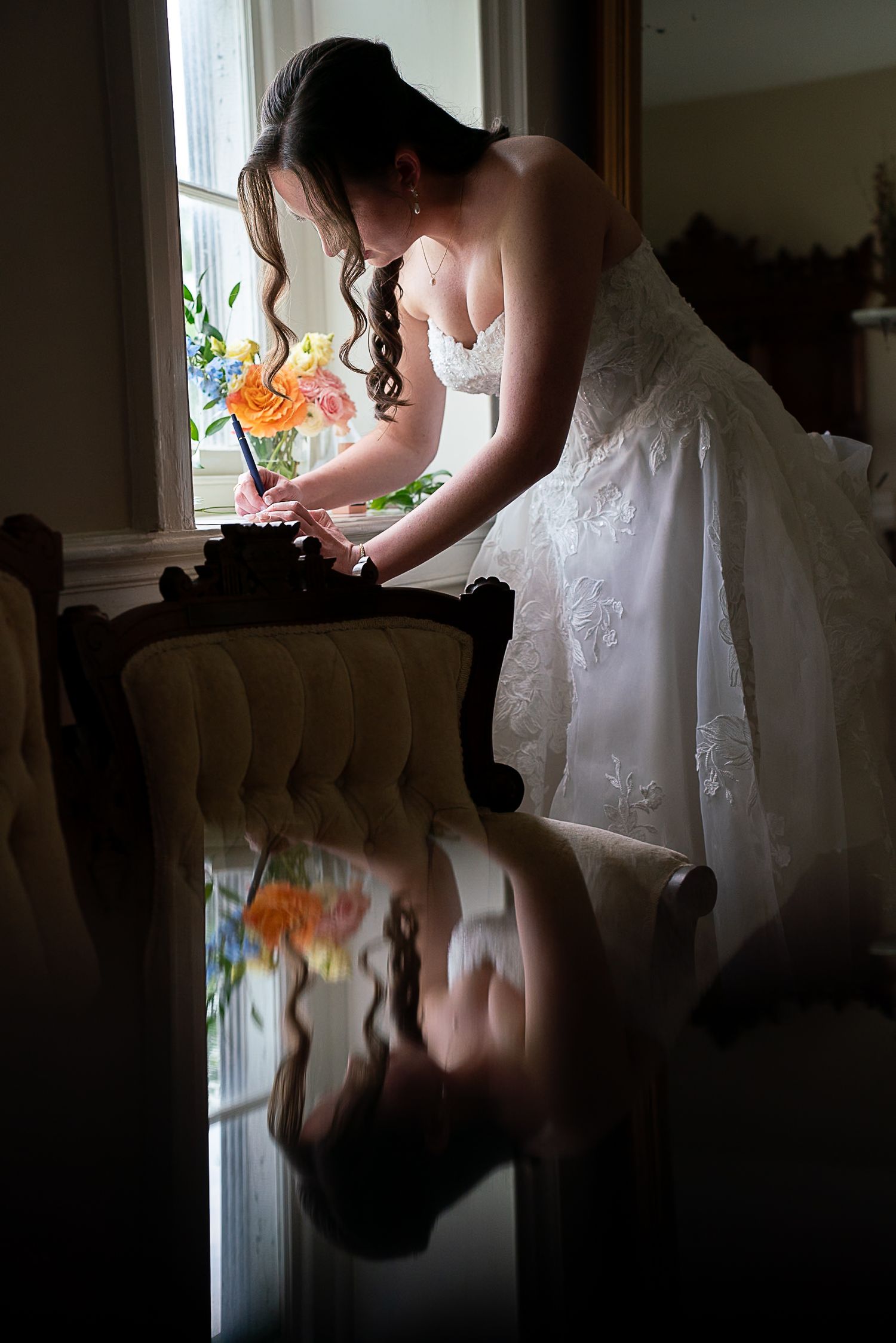 Bride in white lace gown typing on vintage typewriter by window, with colorful floral reflection on glossy floor.