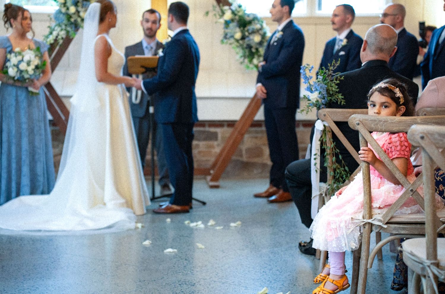 A young flower girl in a pink dress sits on a chair during an elegant wedding ceremony with the bride and groom at the altar.