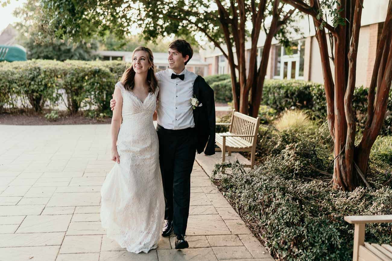 Smiling bride and groom stroll together along a tree-lined stone walkway on their wedding day.