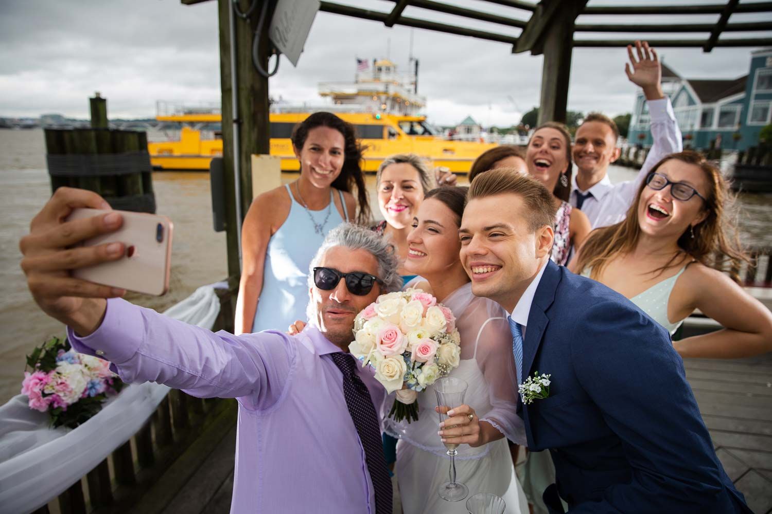 Wedding party takes joyful group selfie with bride holding pink bouquet near waterfront dock.