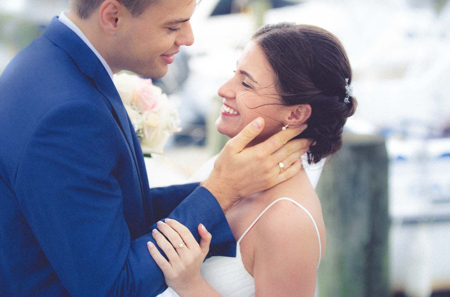 Groom tenderly cupping bride's face as she smiles joyfully at a marina on their wedding day.