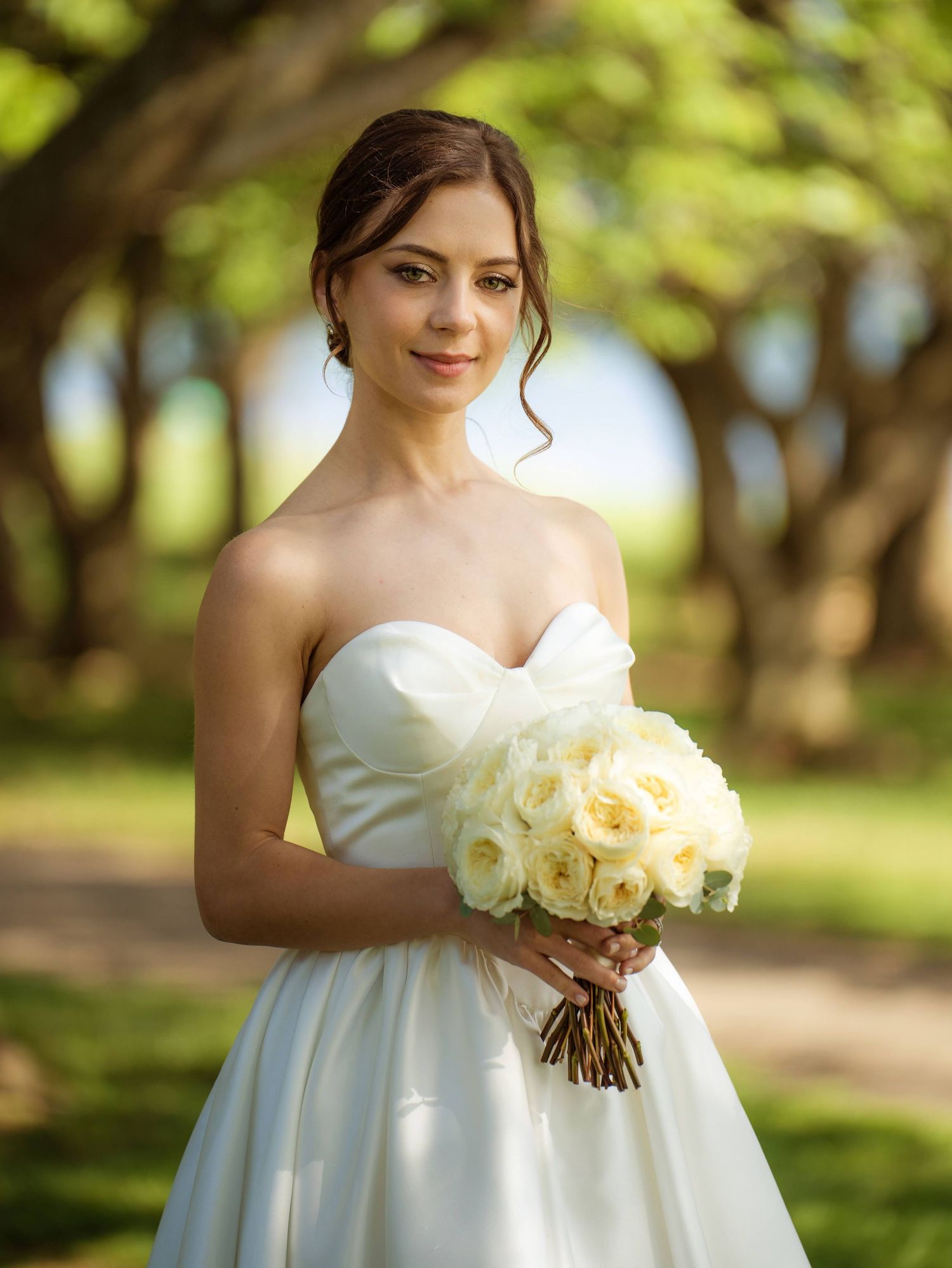 Elegant bride in strapless white gown holds cream rose bouquet among lush green trees outdoors.