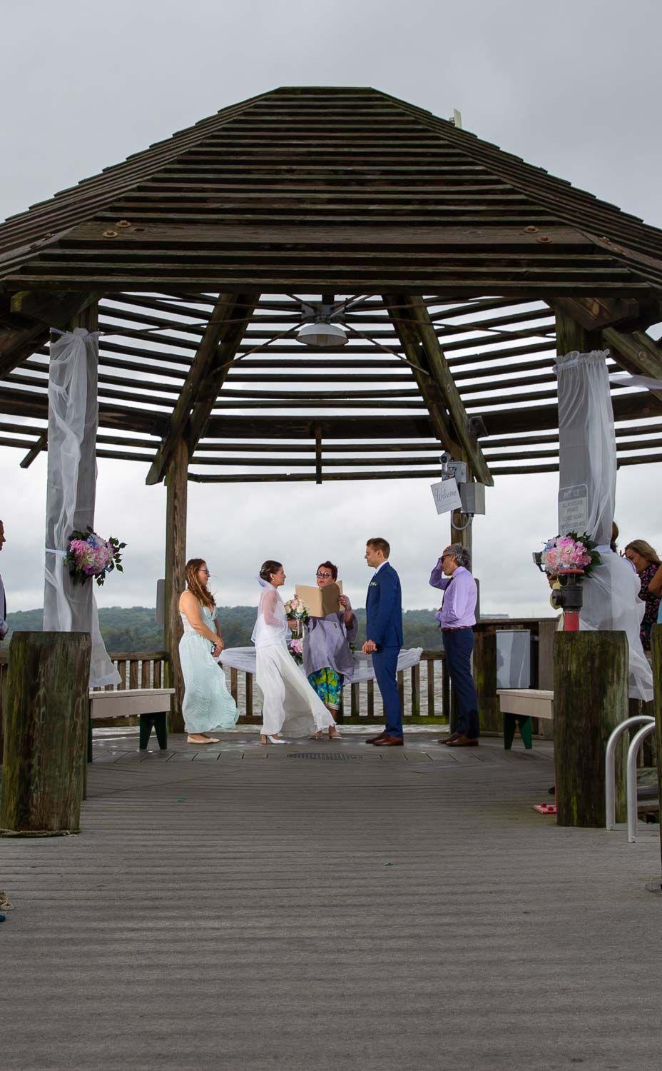Intimate outdoor wedding ceremony under a rustic wooden gazebo on a waterfront pier.