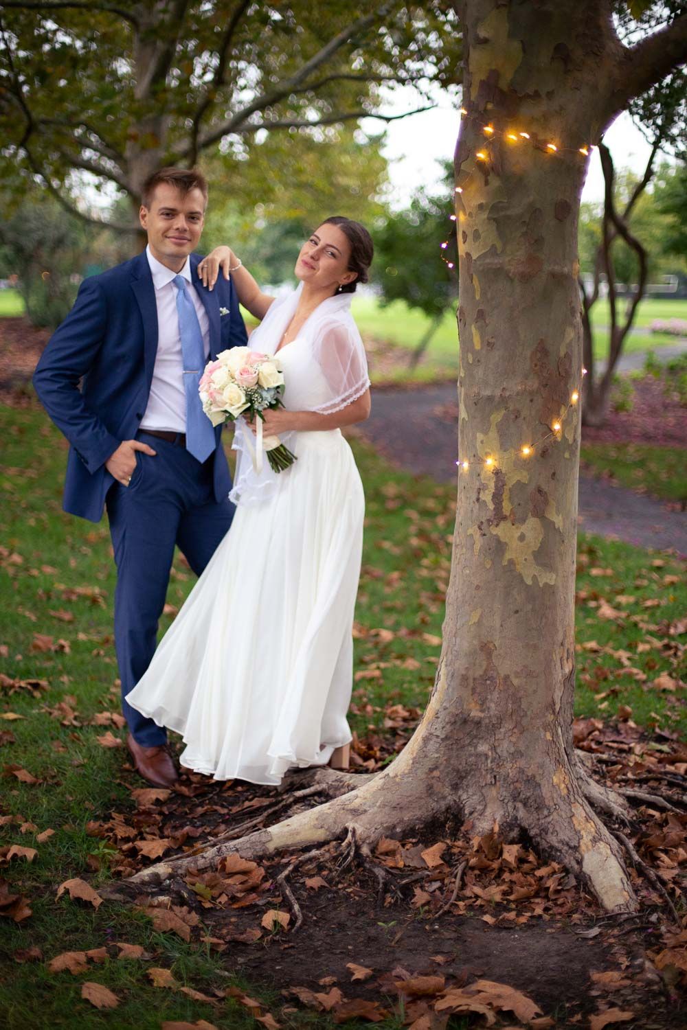 Bride and groom posing together by a tree with fairy lights surrounded by autumn leaves in a park.
