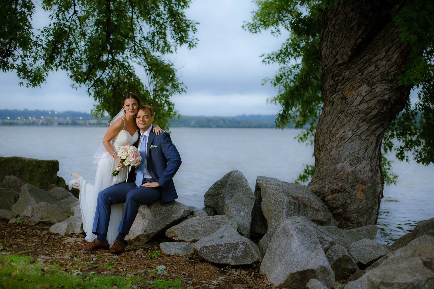 Bride and groom sitting on rocks under a large tree by a lake on their wedding day.