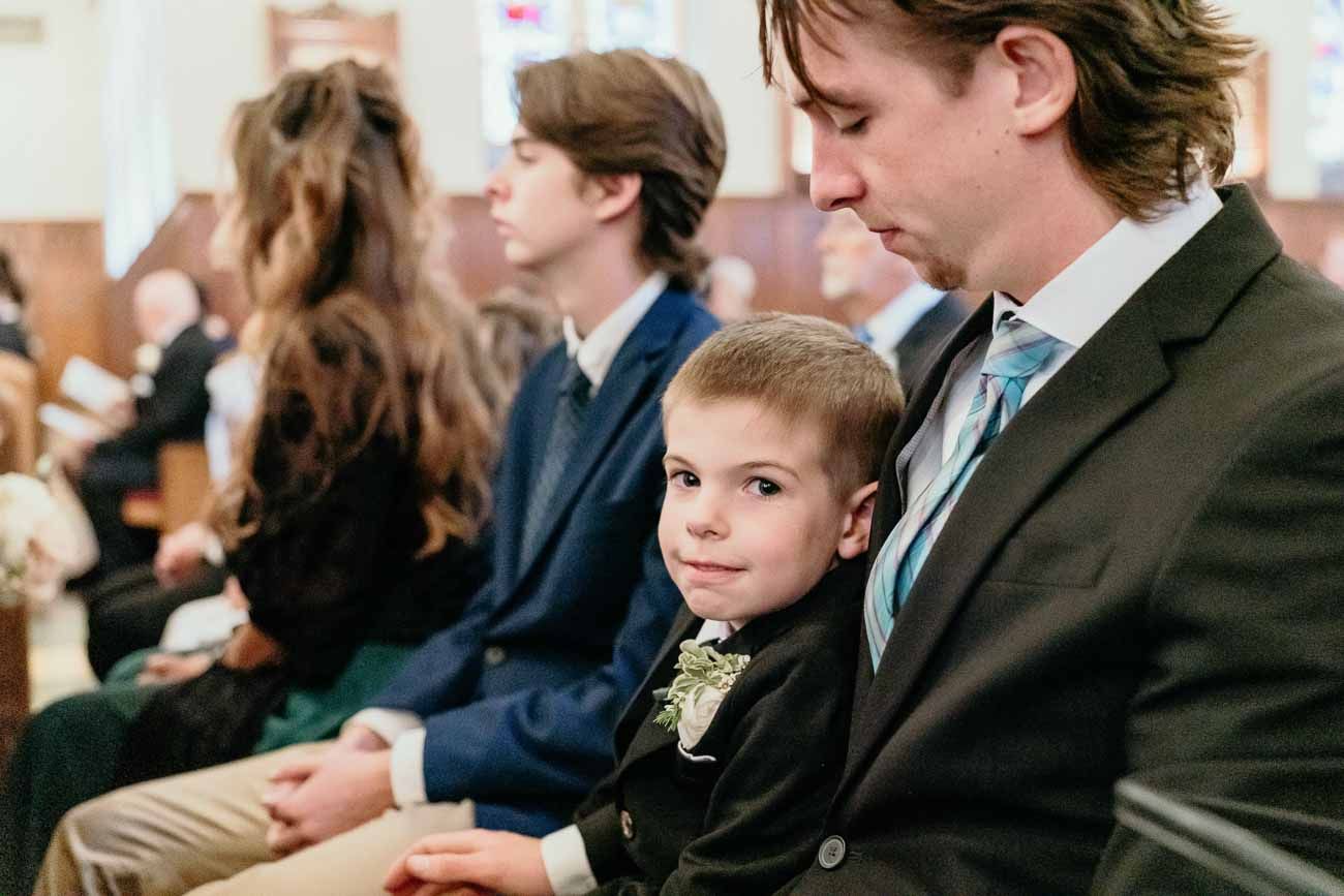 Young boy in suit leans against father while seated in church pew during wedding ceremony.