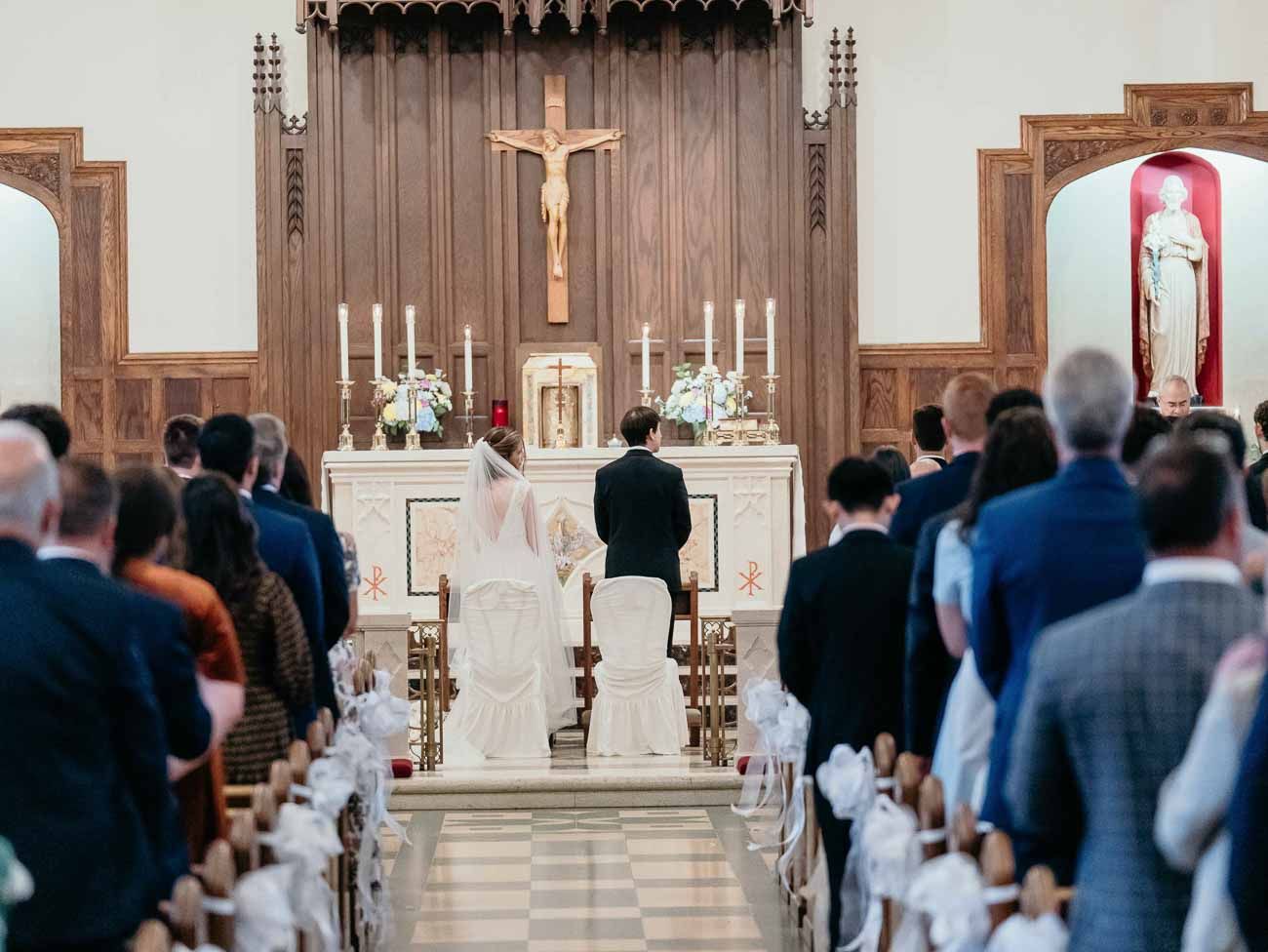Bride and groom kneeling at the altar during a Catholic wedding ceremony with guests standing in pews.