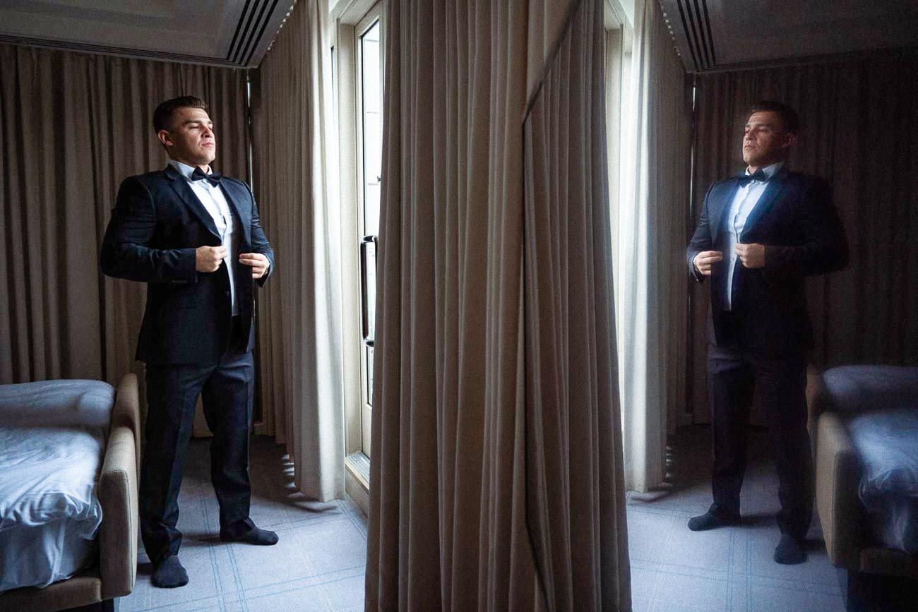 Groom in navy tuxedo adjusting bow tie reflected in full-length mirror.