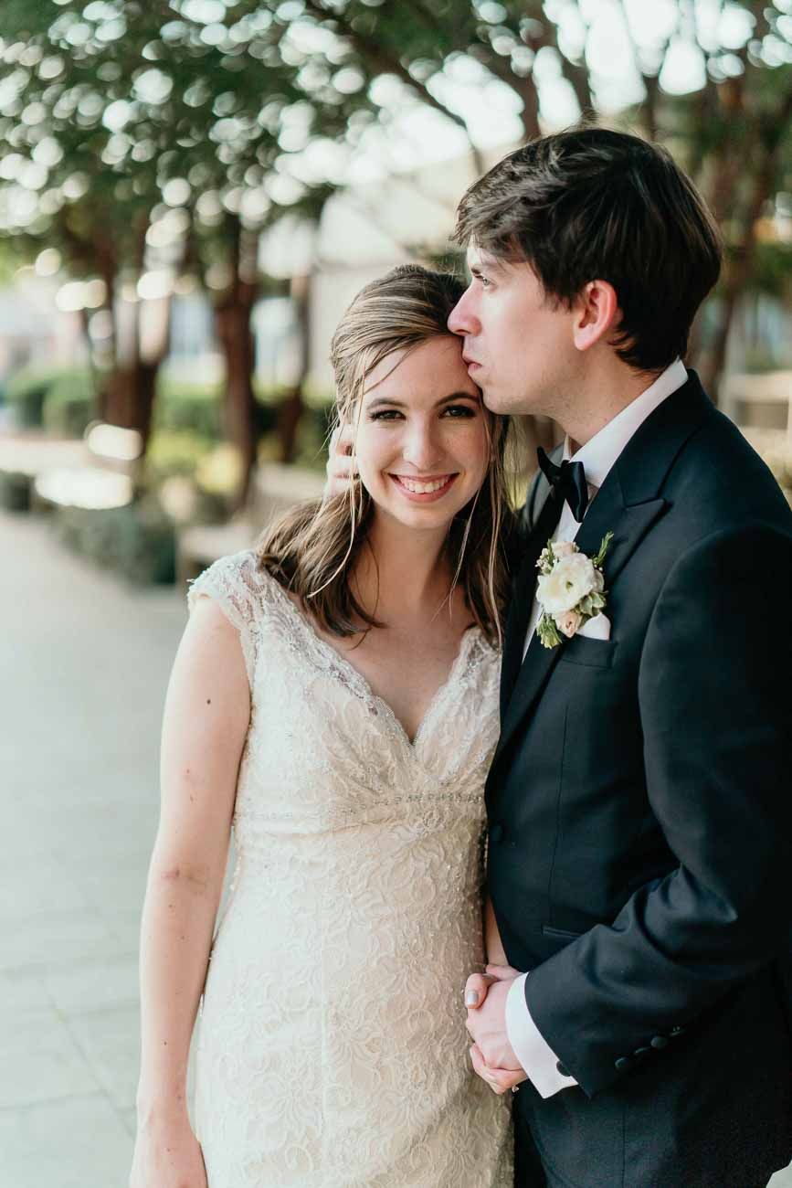 Groom in black suit kissing bride on the cheek as she smiles, standing on a tree-lined walkway.
