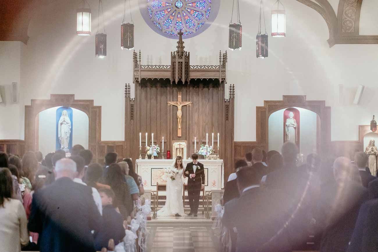 Wide view of Catholic church wedding ceremony with guests seated in pews and couple at the altar.