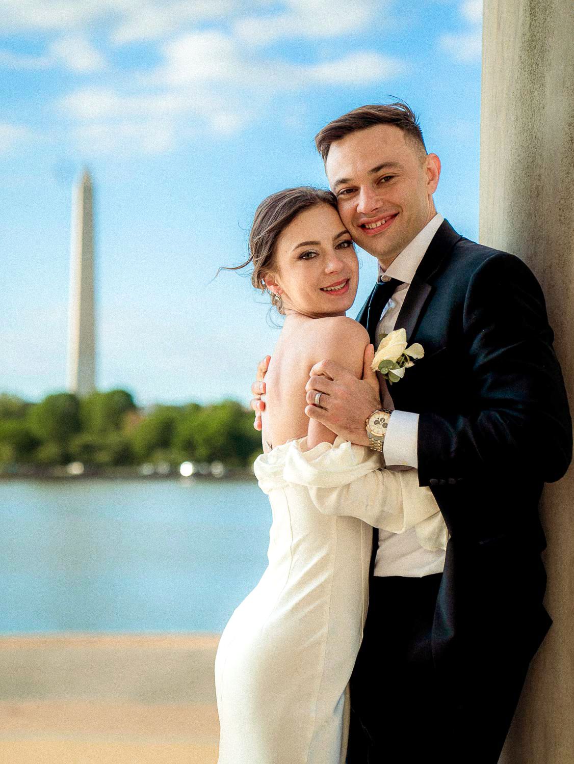 Smiling newlyweds pose together against marble column at Jefferson Memorial with Washington Monument in background.