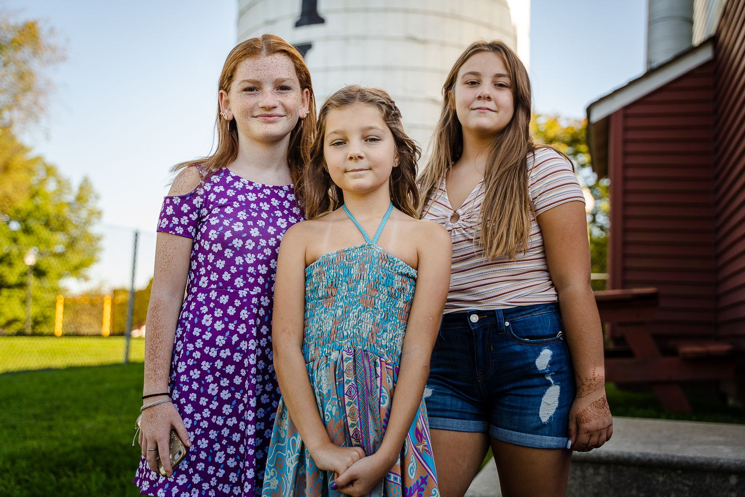 Three girls posing outdoors near a white silo on a sunny day.