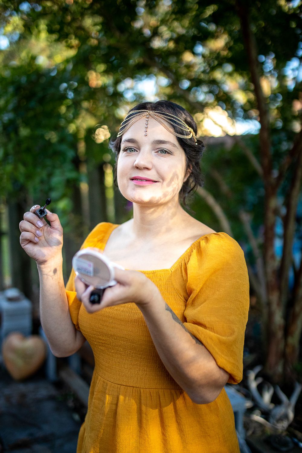 Smiling woman in yellow dress with gold headchain applying makeup outdoors in garden.