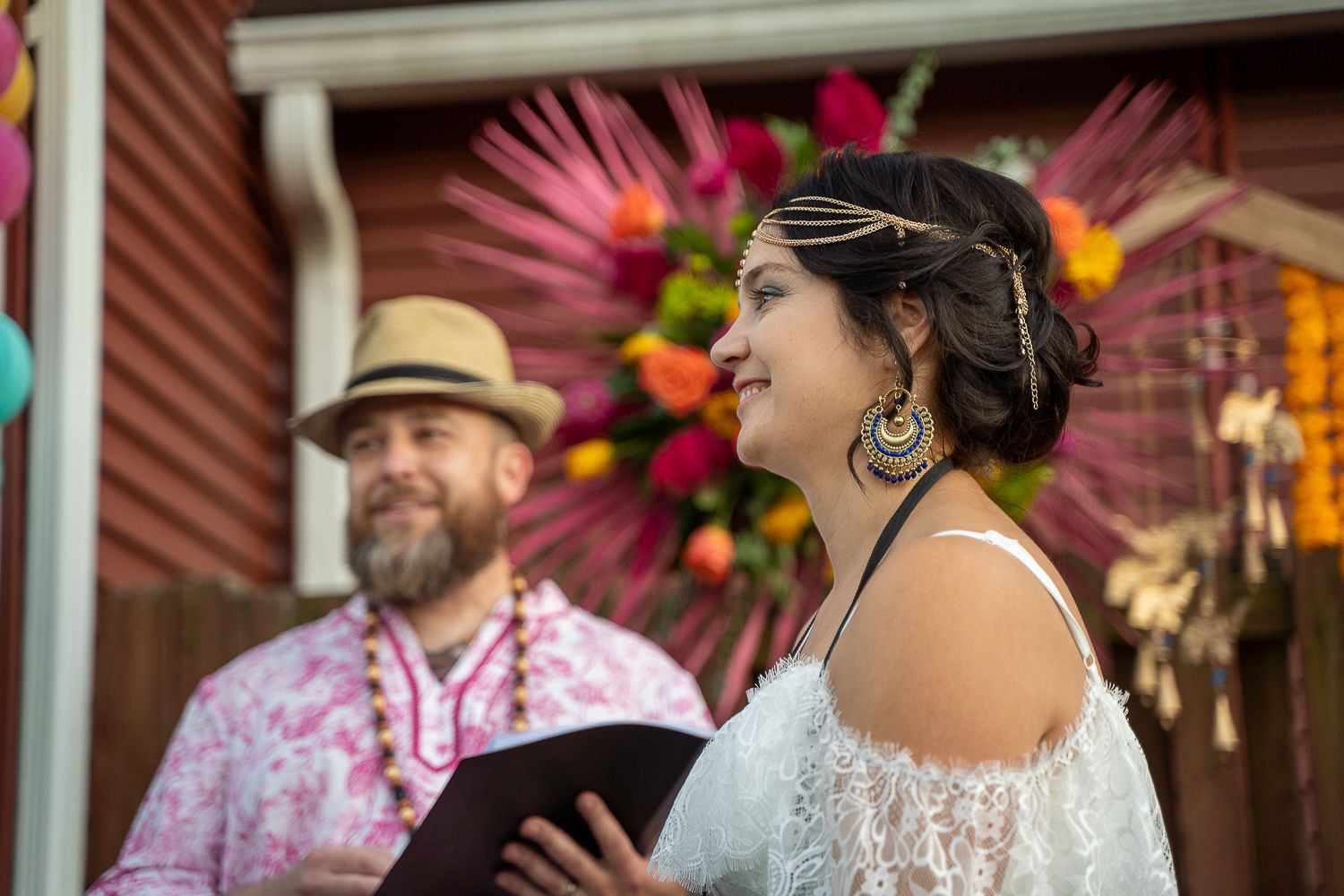 Bride in boho lace dress with gold headpiece reads vows as officiant in pink shirt holds book at colorful outdoor wedding.