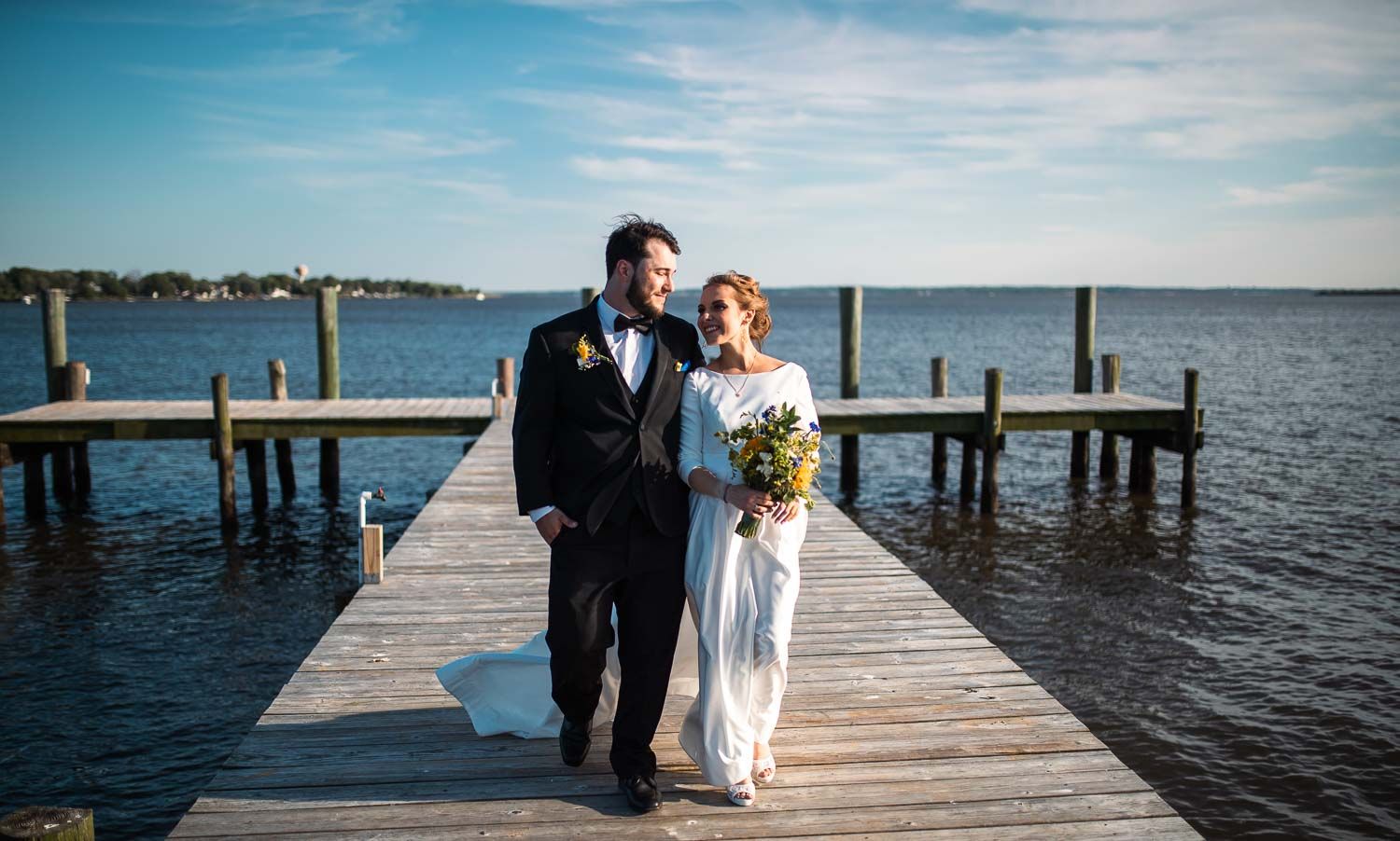 Newlywed couple walking hand in hand on wooden pier over calm blue water on their wedding day.