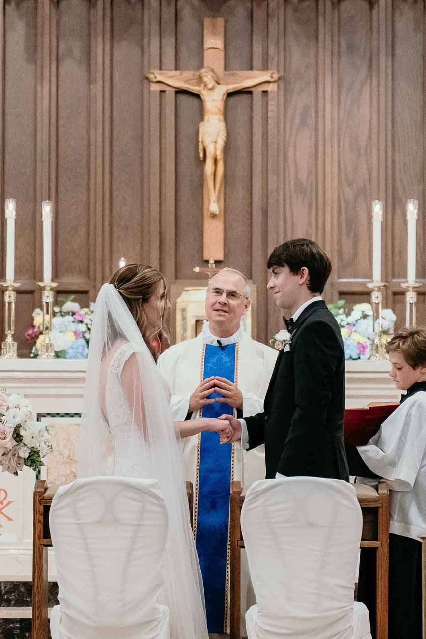 Bride and groom holding hands before priest during Catholic wedding ceremony under crucifix.
