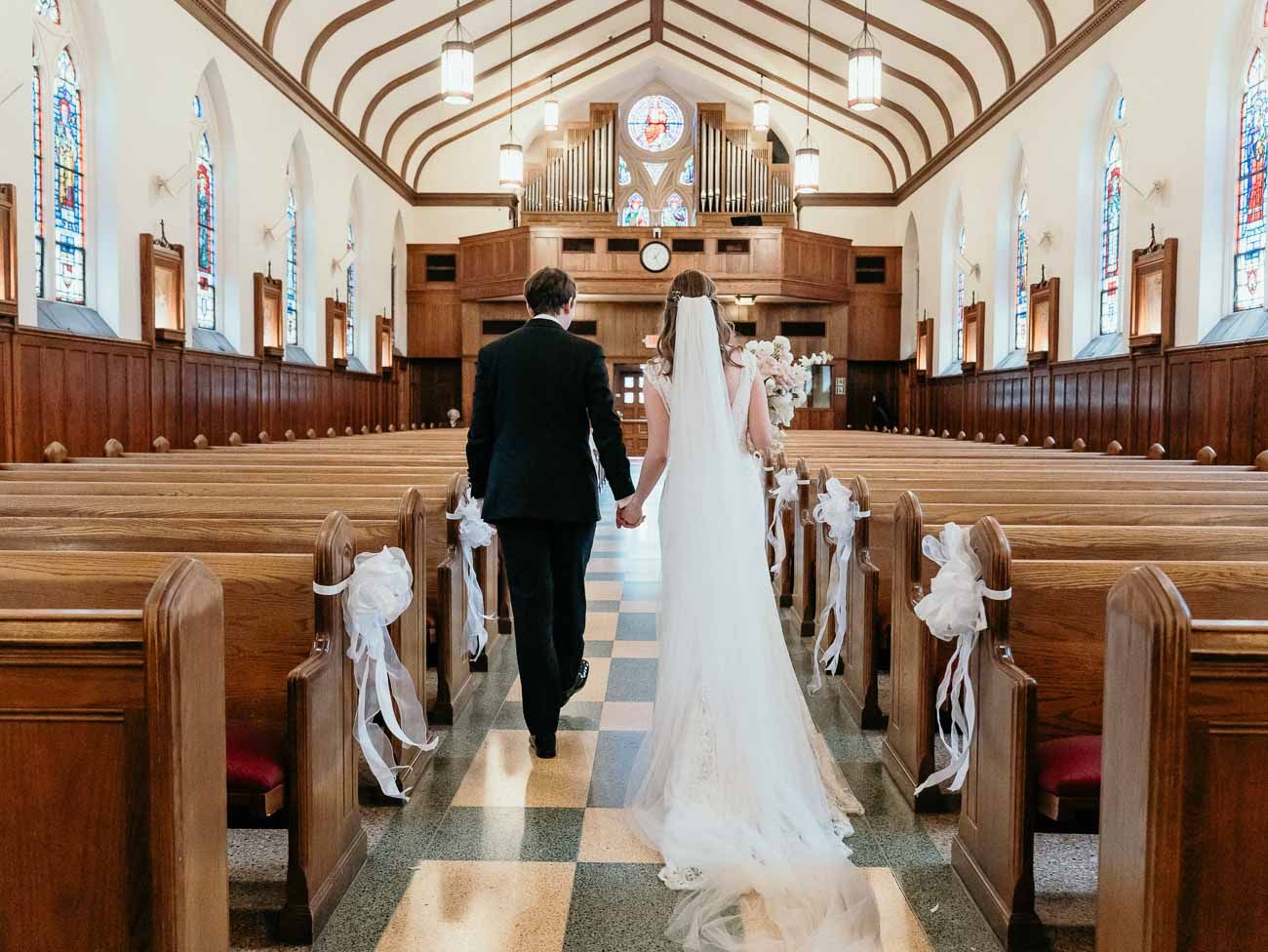 Bride and groom walk hand in hand down a sunlit church aisle adorned with white ribbon bows.