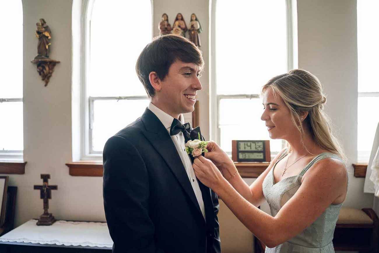 Bridesmaid pins a boutonniere on the smiling groom inside a church before the wedding ceremony.