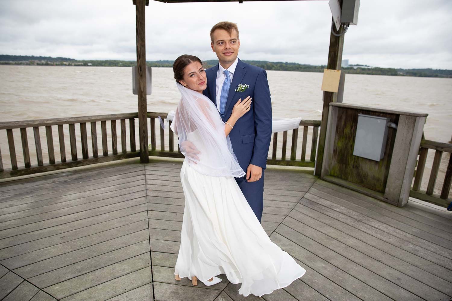 Happy newlyweds pose together on wooden deck overlooking river with bride in flowing white gown.