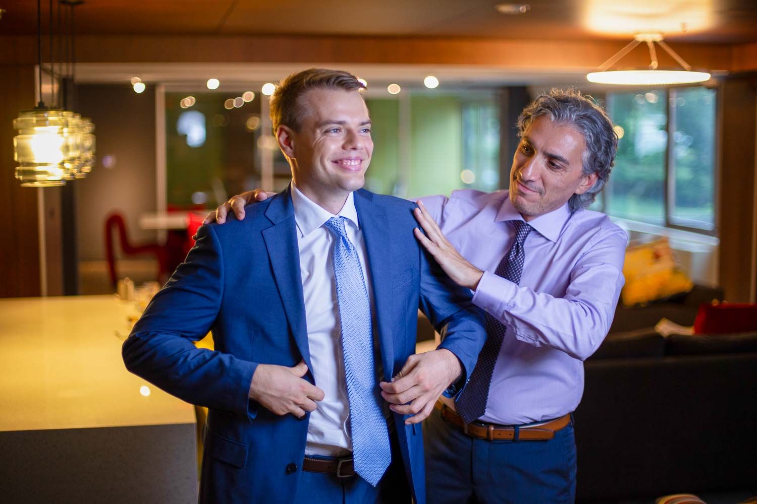 Older man helping a young groom adjust his blue suit jacket in a warmly lit modern venue.