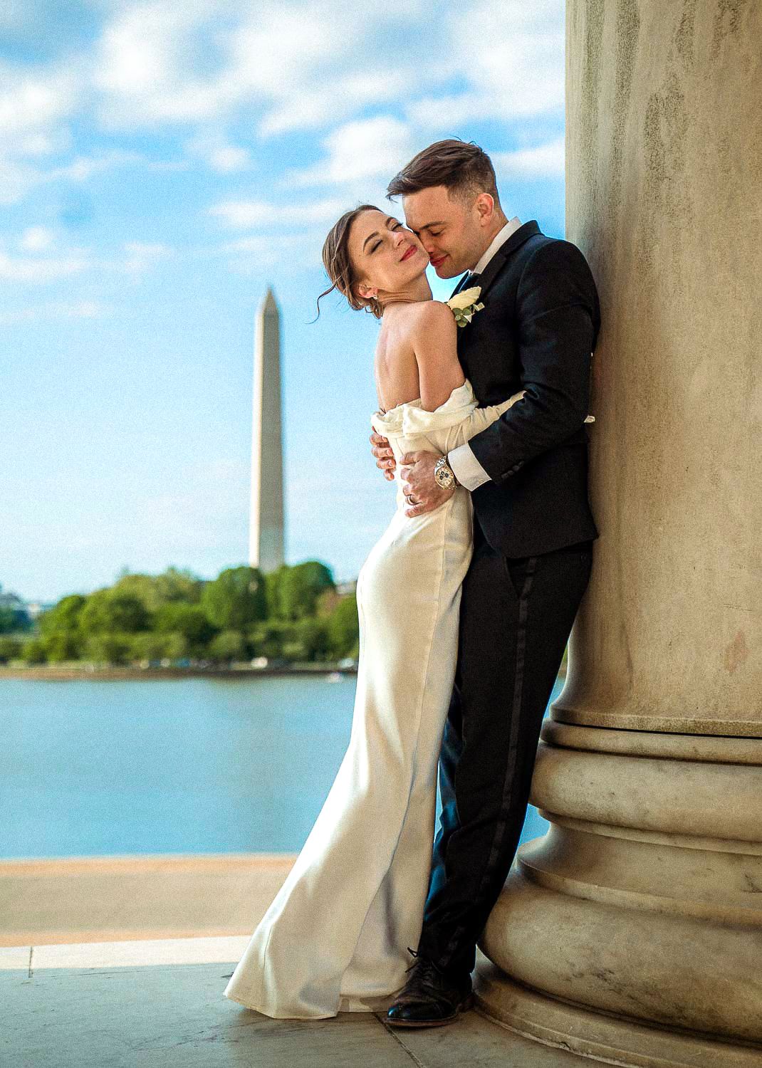 Wedding couple embraces by a marble column with the Washington Monument in the background.