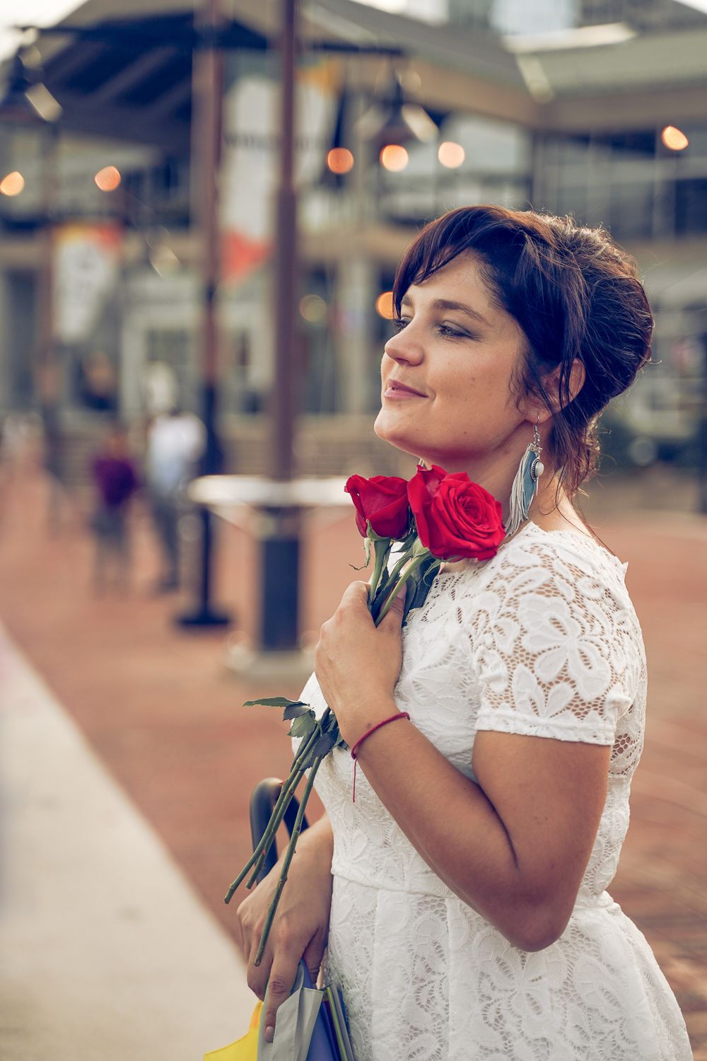 Bride in white lace dress holding red roses smiling softly at outdoor urban wedding venue.