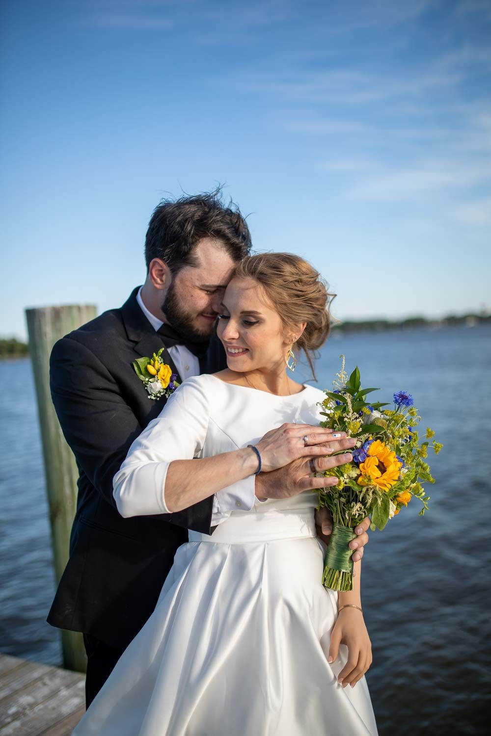 Newlyweds embrace on a dock by the water, bride holding a vibrant sunflower and wildflower bouquet.
