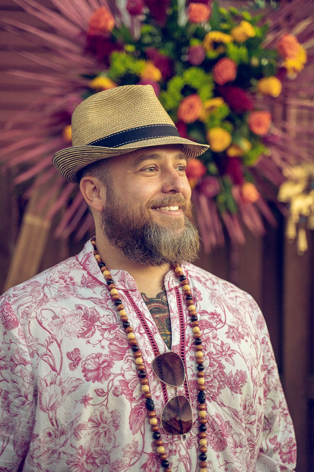 Smiling bearded man in straw fedora and floral shirt with beaded necklace at tropical wedding.
