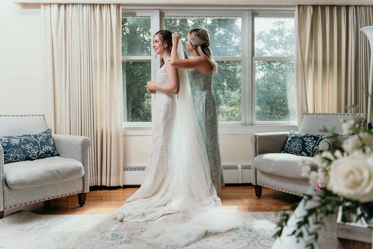 Bridesmaid helps bride with veil in sunlit room with white floral arrangements nearby.