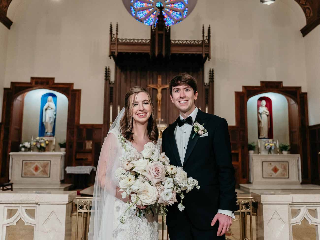 Bride and groom portrait at Catholic church altar with rose window and religious statues behind them.