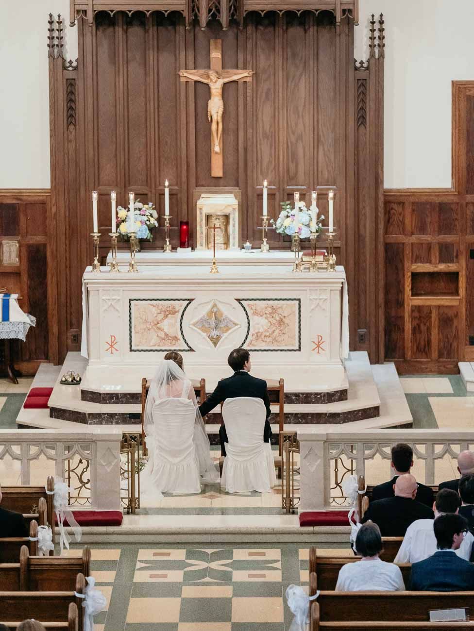 Bride and groom seated before a wooden crucifix altar during a Catholic wedding Mass ceremony.