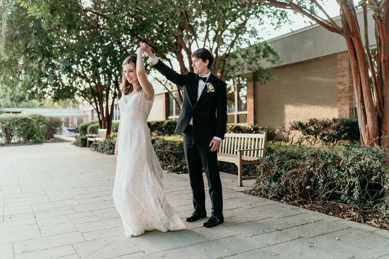Groom spinning bride in lace wedding gown on a sunlit outdoor walkway surrounded by trees.