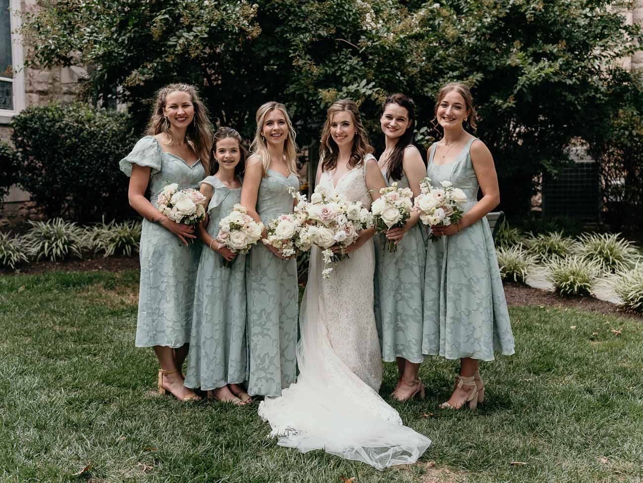 Bride and five bridesmaids in matching sage green floral dresses pose with white floral bouquets.