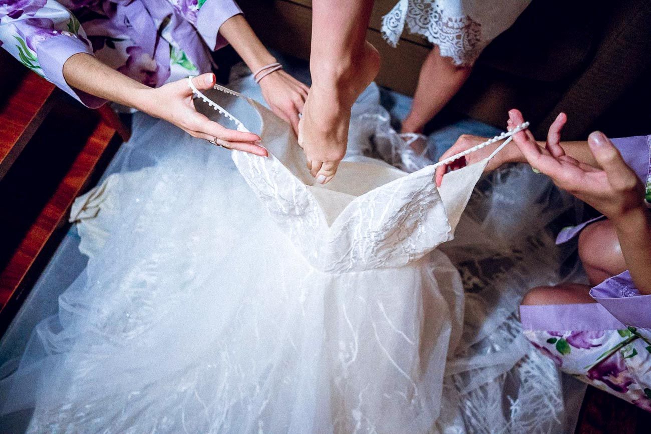 Bridesmaids helping bride lace up elegant white wedding gown with pearl details from above.