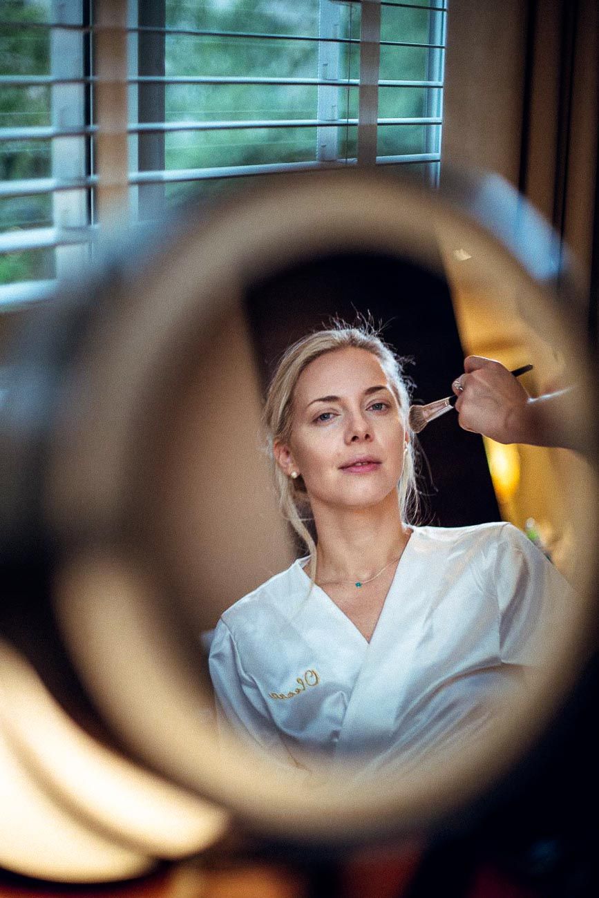 Blonde bride in white robe getting makeup applied reflected in round mirror.