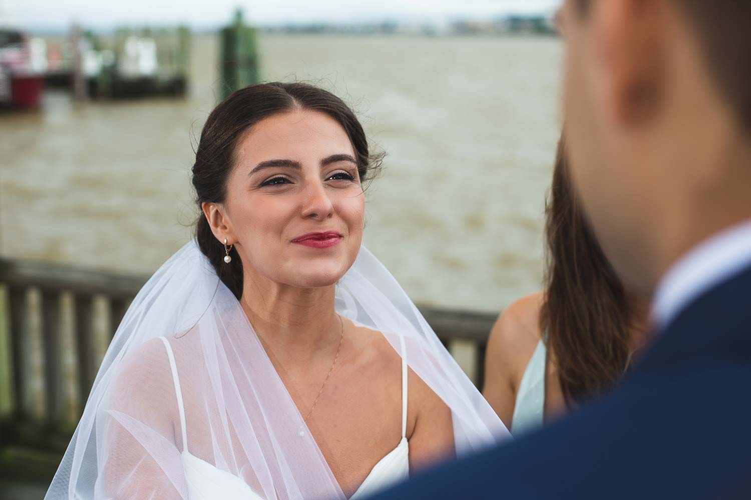 Beautiful bride in white veil gazes lovingly at groom during outdoor waterfront wedding ceremony.