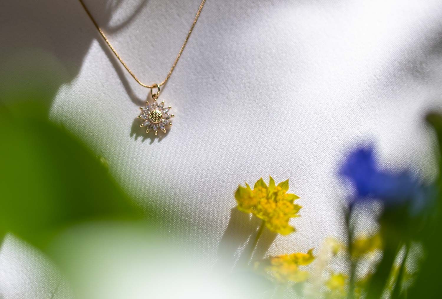 Delicate diamond sunflower pendant necklace on gold chain displayed against white fabric with yellow and blue flowers.