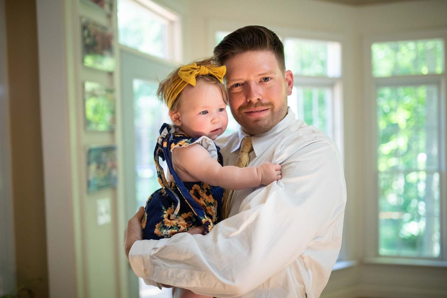 Proud father in white dress shirt holding baby girl with yellow bow at indoor event.