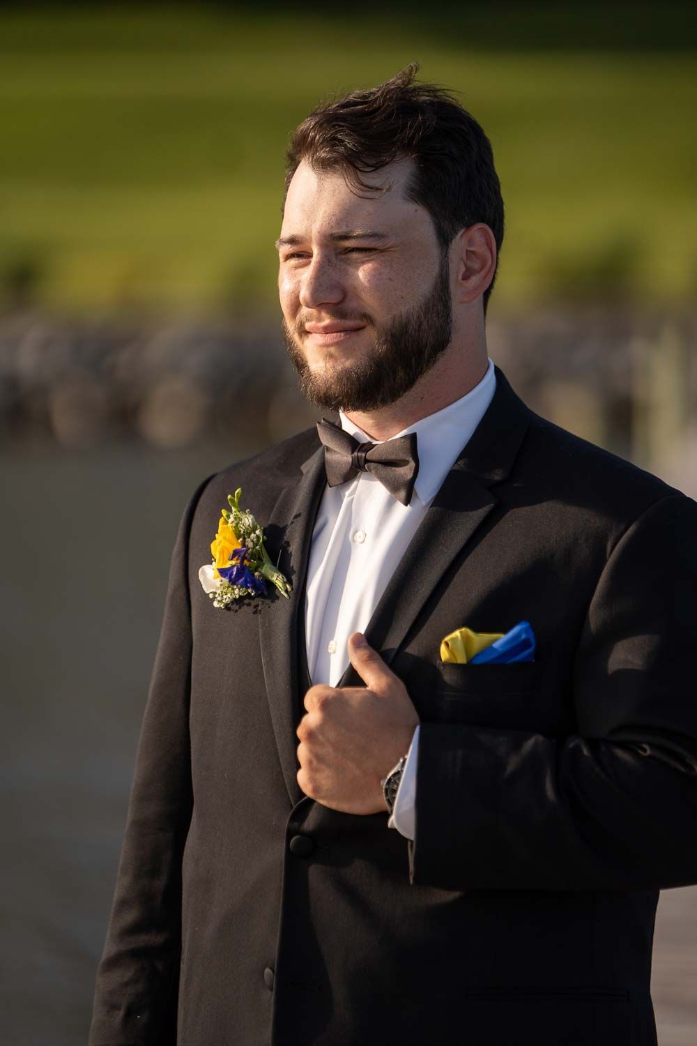 Handsome groom in a black tuxedo with boutonniere and yellow pocket square smiling outdoors.