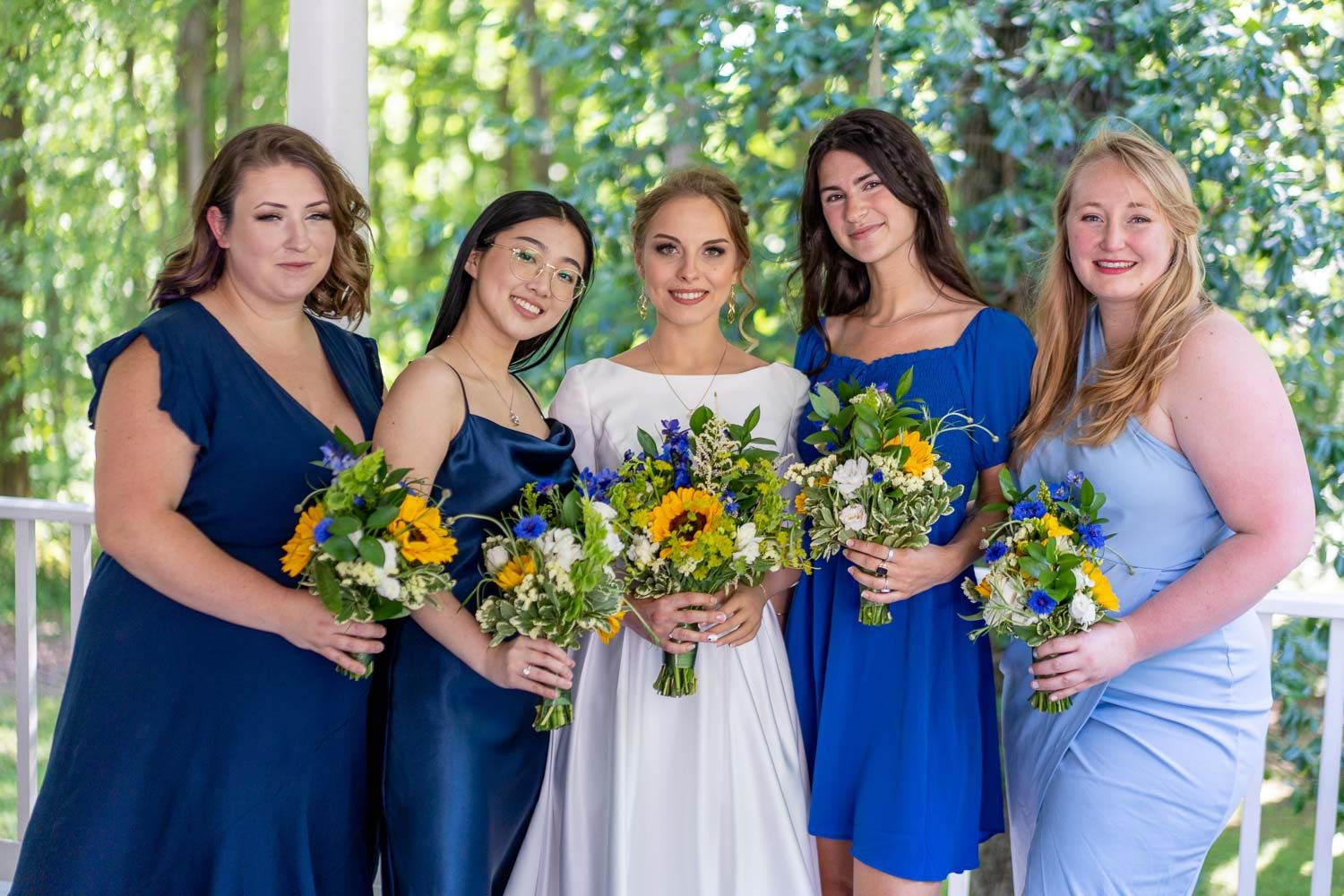 Bride with four bridesmaids in blue dresses holding sunflower bouquets under a white gazebo.