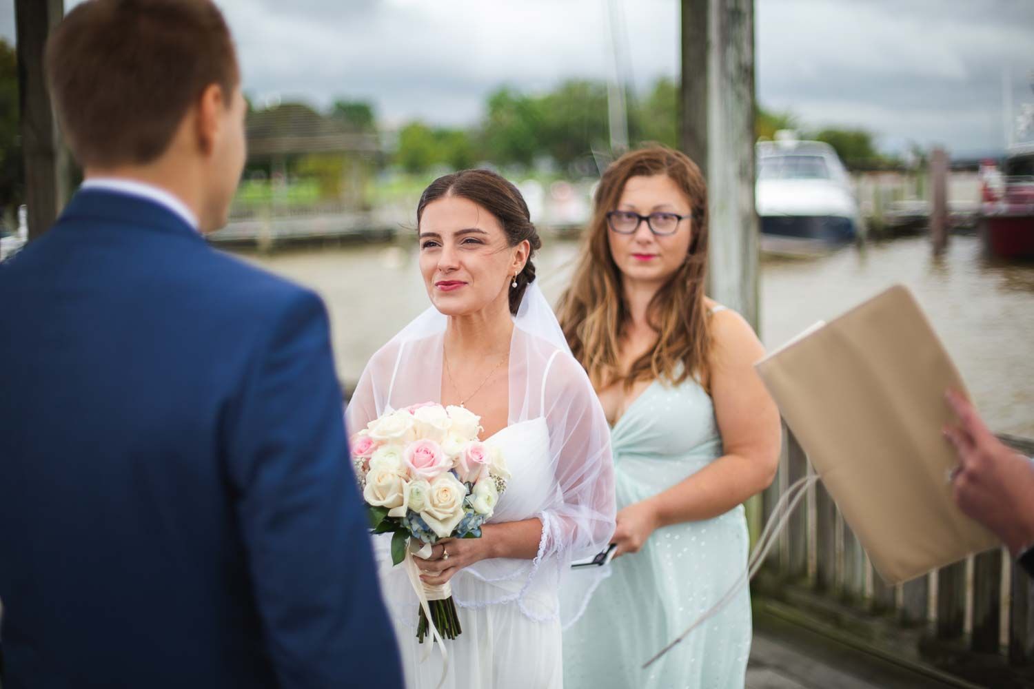 Bride in white veil holds pink bouquet while bridesmaid in mint dress stands beside her at waterfront ceremony.
