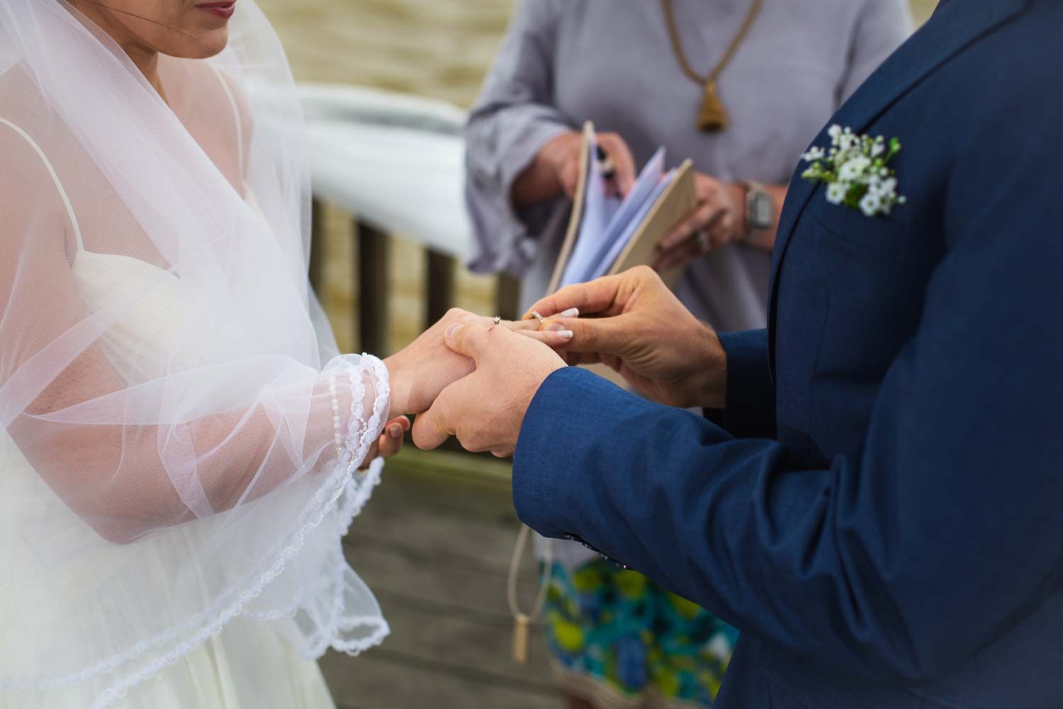 Groom places wedding ring on brides finger as officiant reads from book during intimate outdoor ceremony.