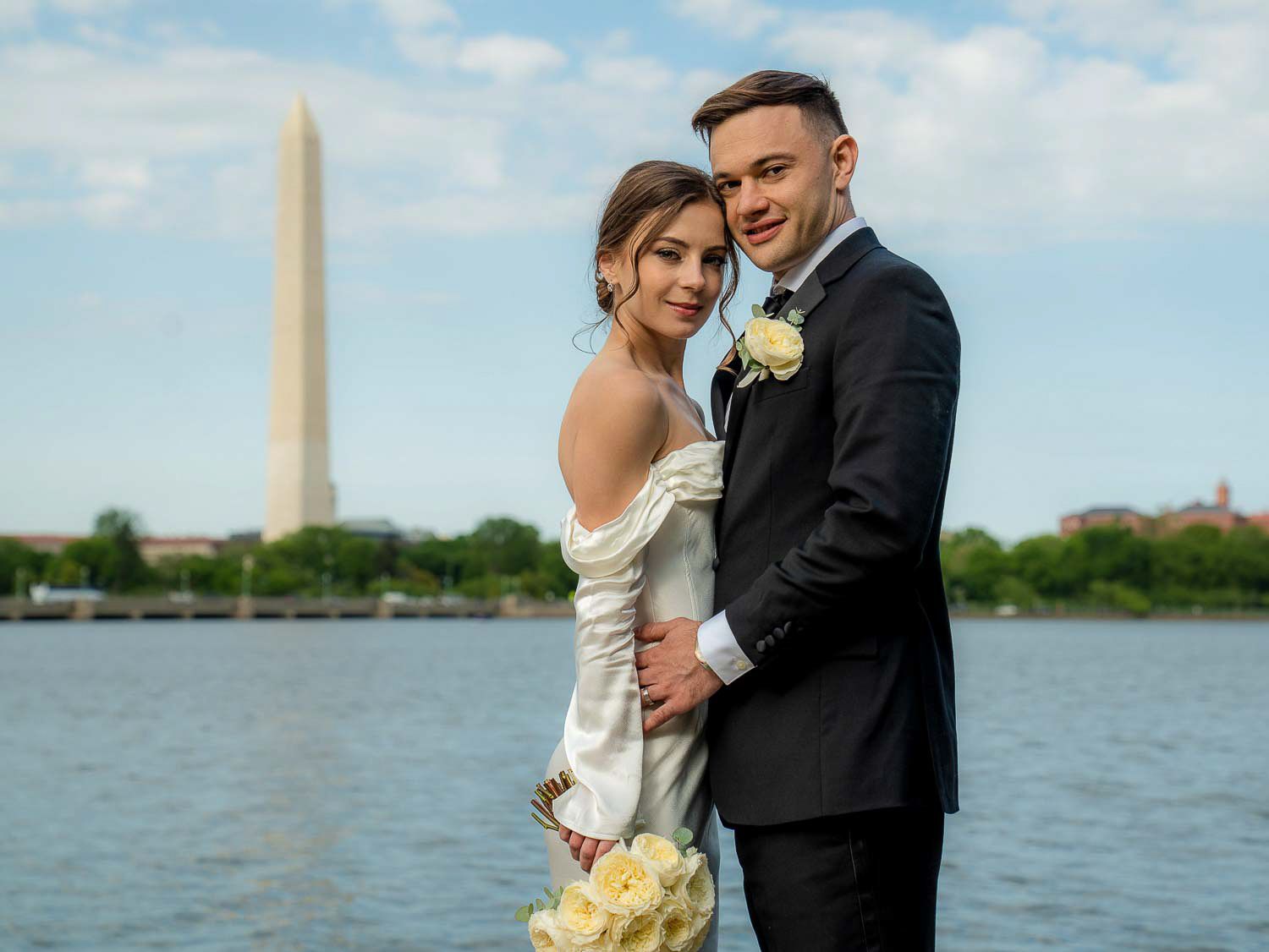 Bride and groom pose by Tidal Basin with Washington Monument visible across the water on sunny day.