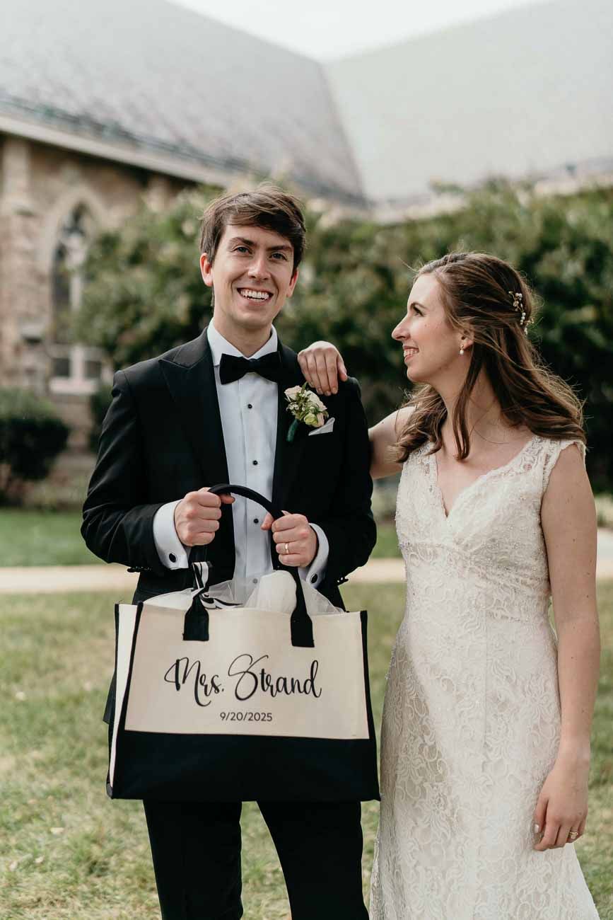 Smiling groom holds a Mrs. Brand tote bag beside his bride in a lace wedding gown outdoors.