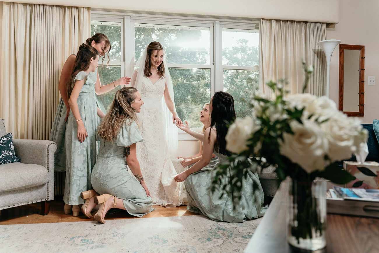 Bridesmaids in sage green dresses help bride adjust white lace wedding gown in bright room.
