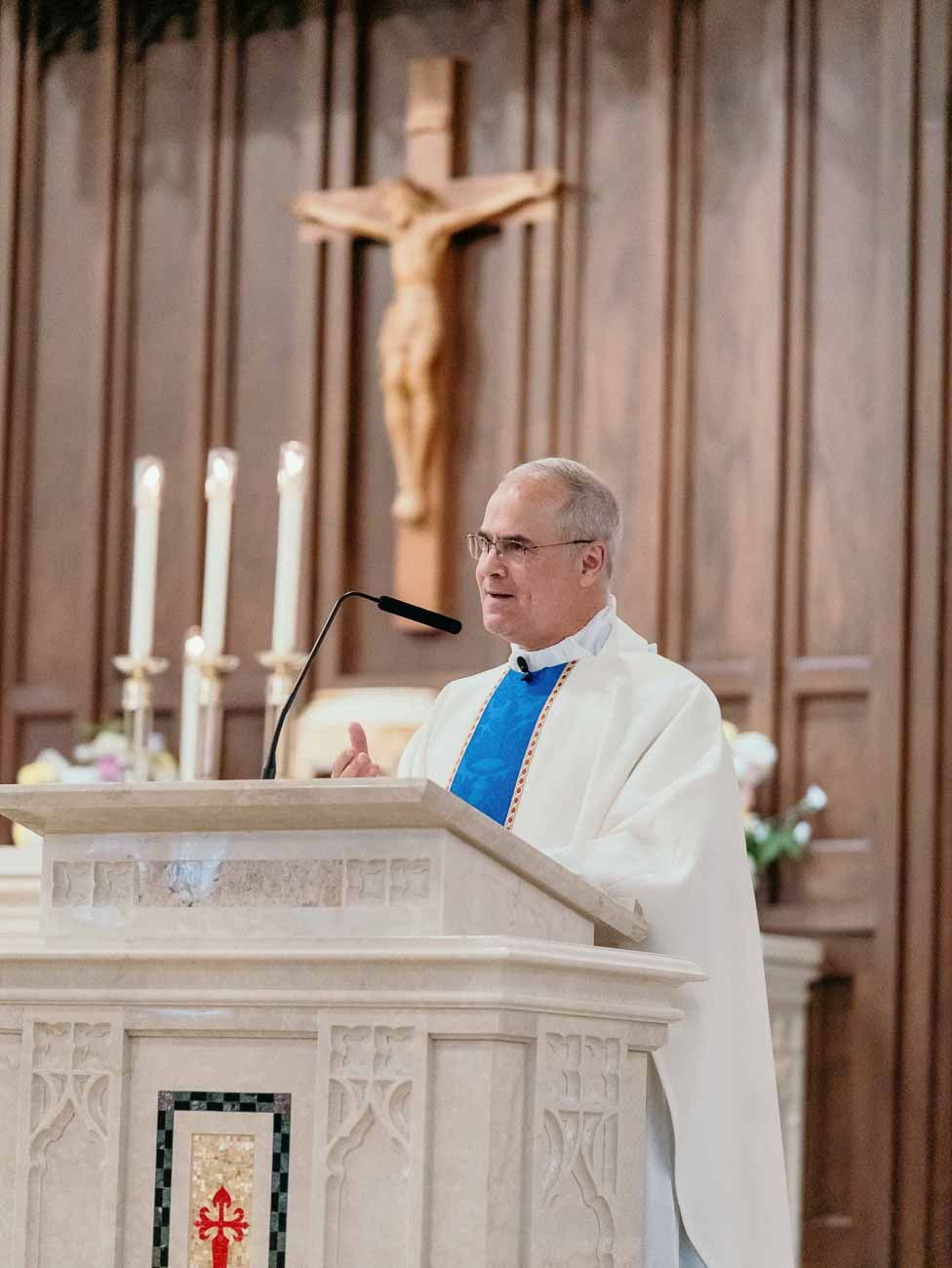 Catholic priest in white vestments delivering a homily at a marble pulpit during a wedding Mass.